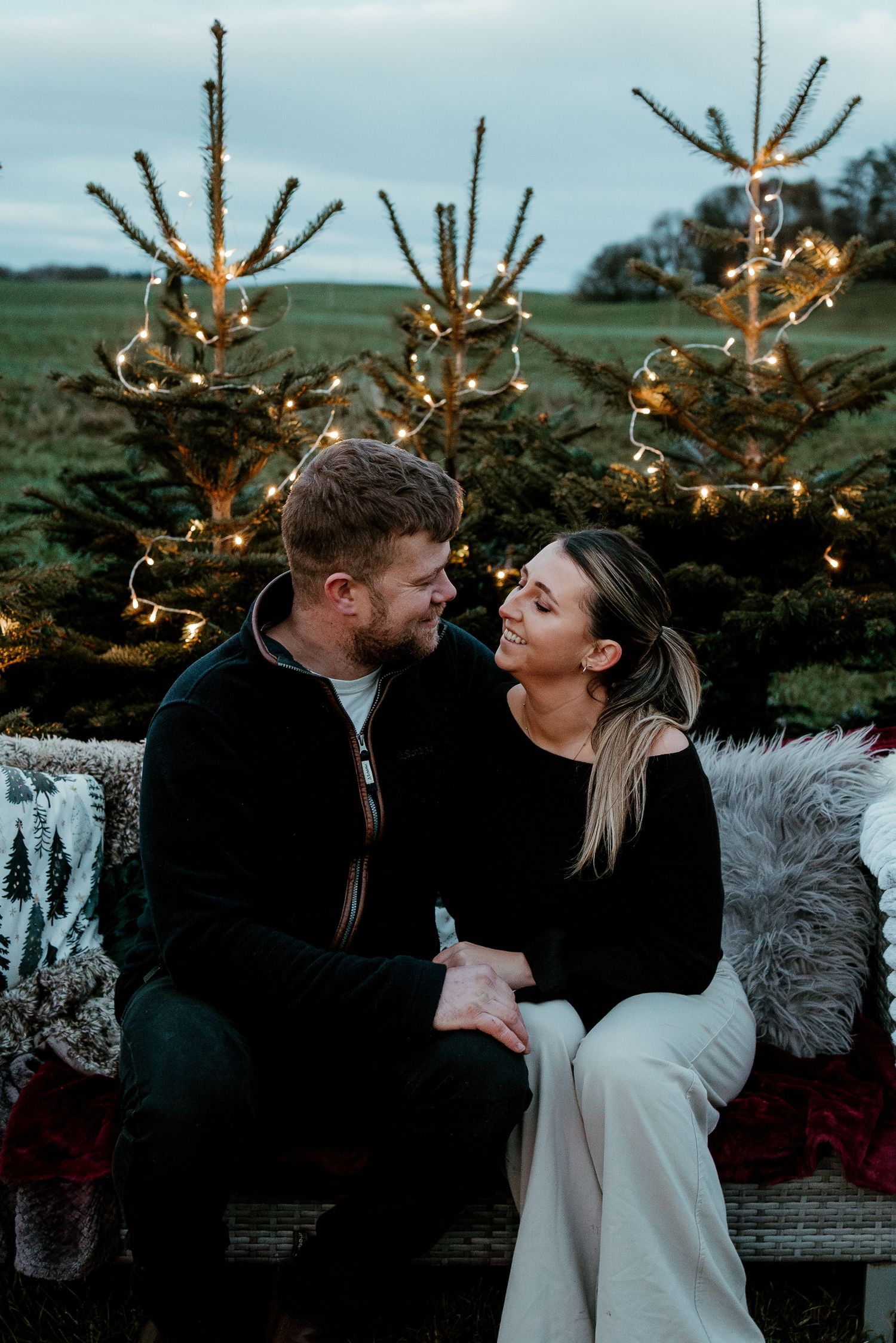 Romantic winter scene with string lights and pine trees as couple shares intimate moment on bench.