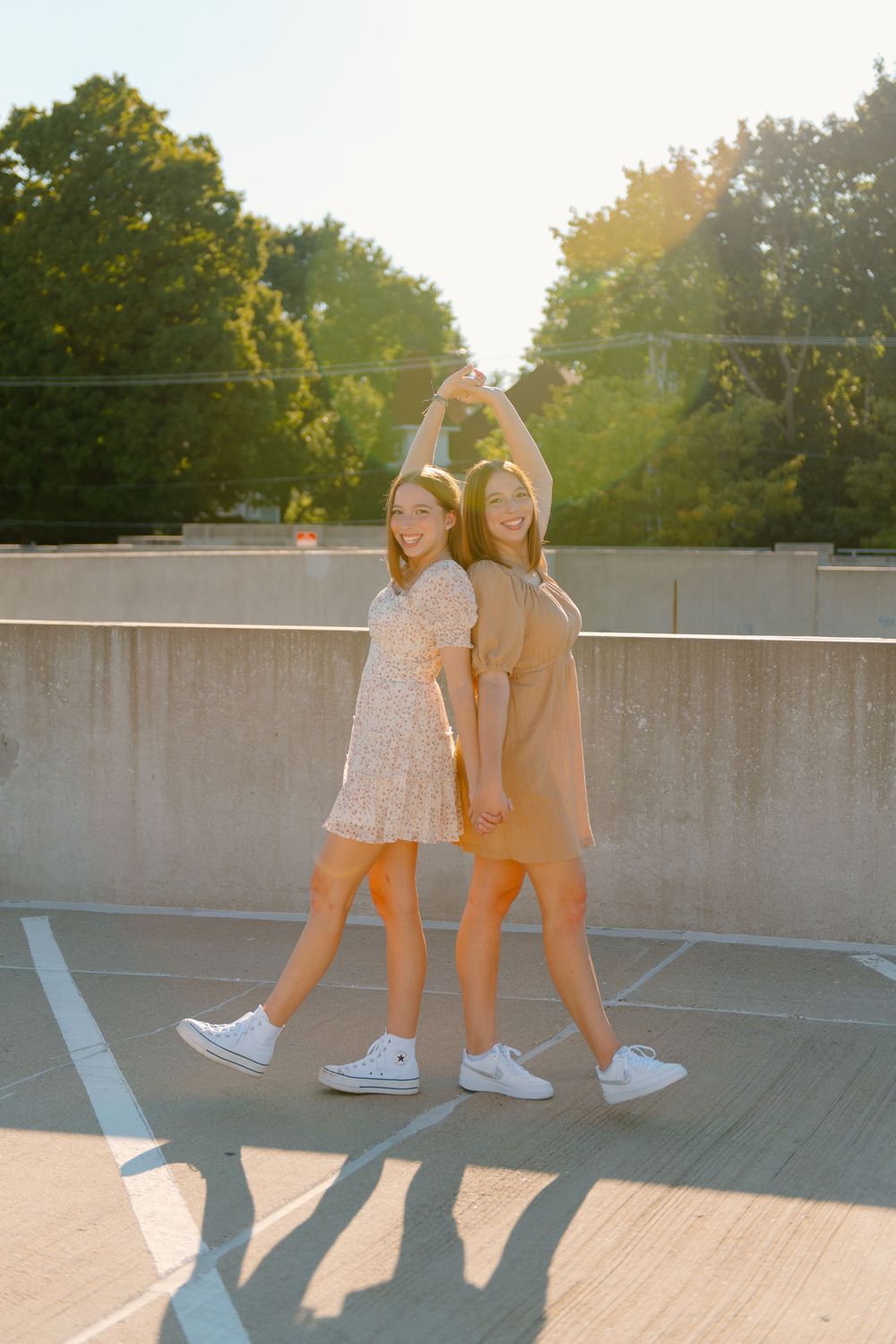 Two best graduate friends in white shoes pose together on a rooftop parking structure at sunset creating long shadows.