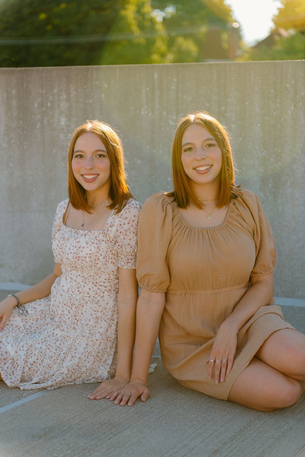 Two graduate women in summer dresses sit together on concrete during golden hour sunshine.