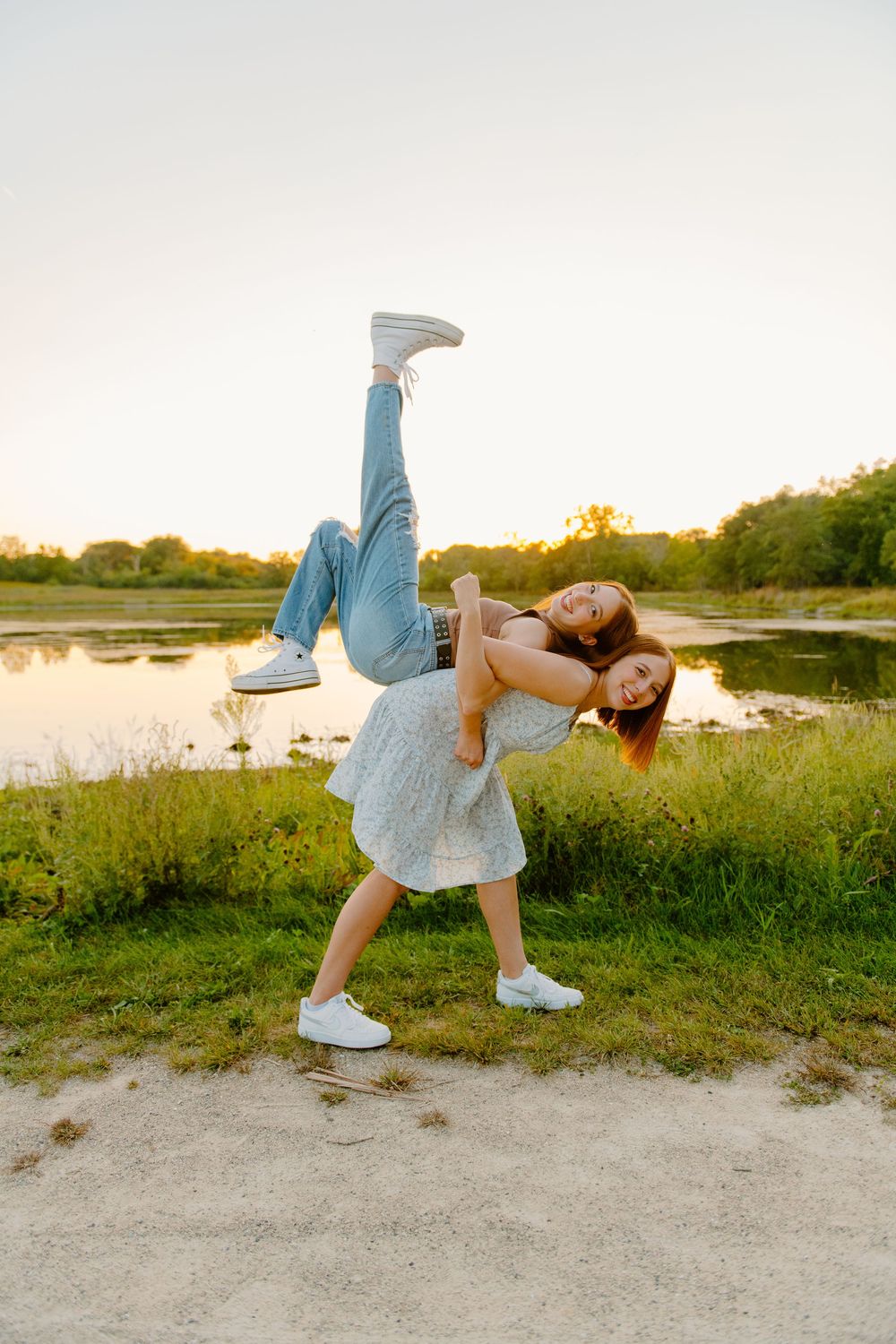 Two graduate sisters having fun outdoors by a lake at sunset, with one playfully lifting the other in a dramatic pose.