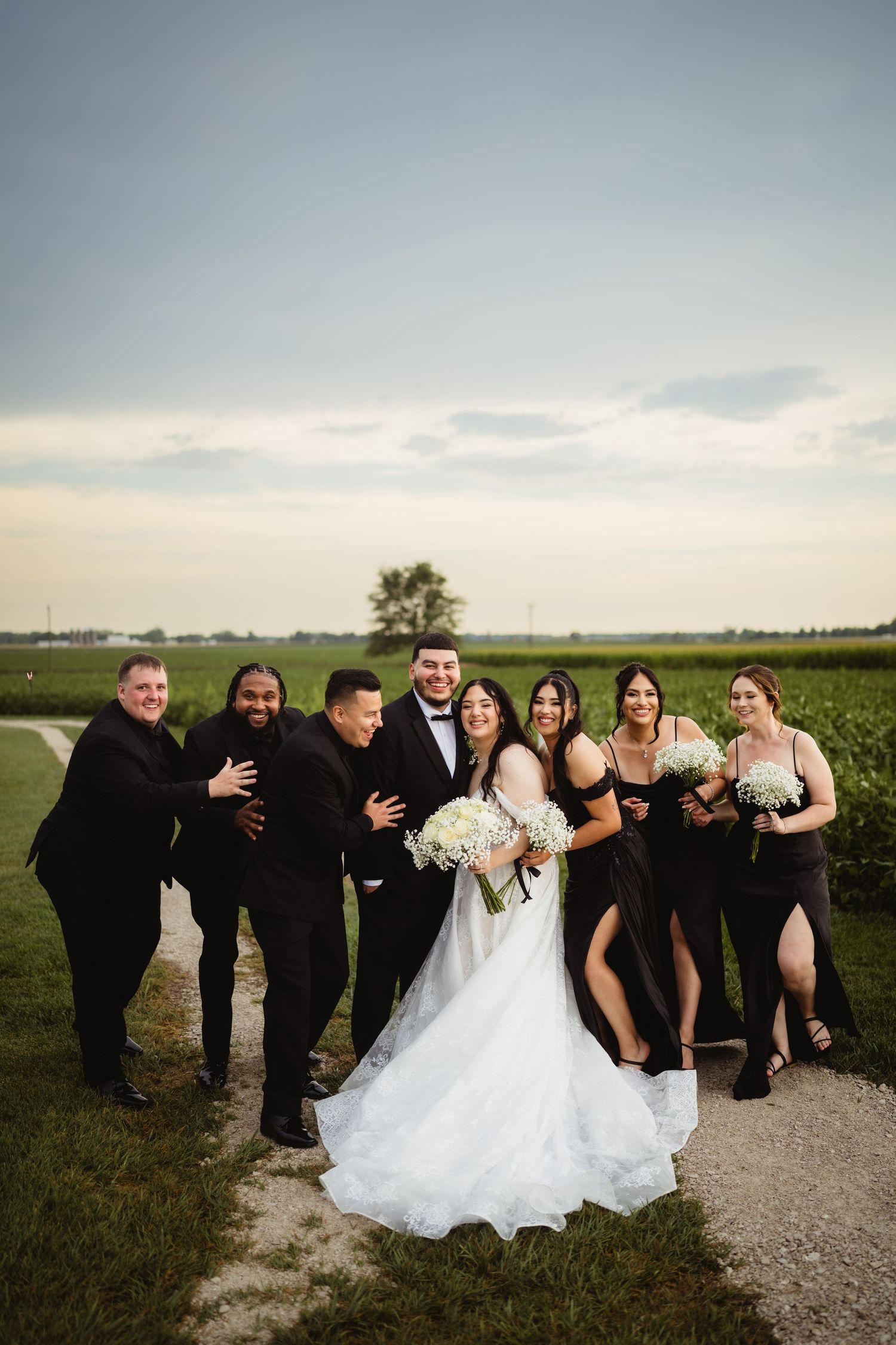 A wedding party in black and white attire poses together on a dirt road with green fields and cloudy sky in the background.
