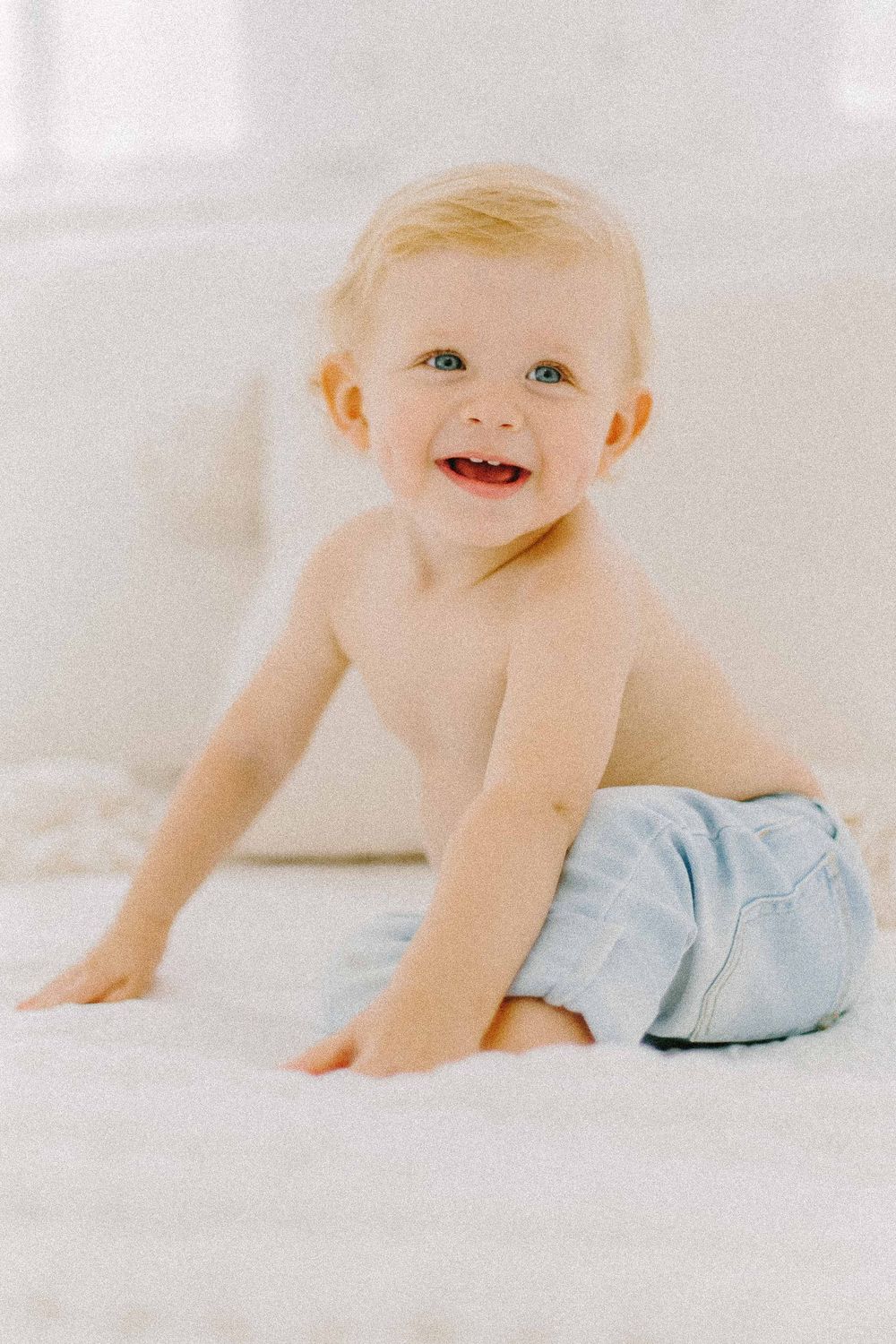 Happy baby in light blue pants crawling on white bedding. Specializing in maternity, newborn, and family photography