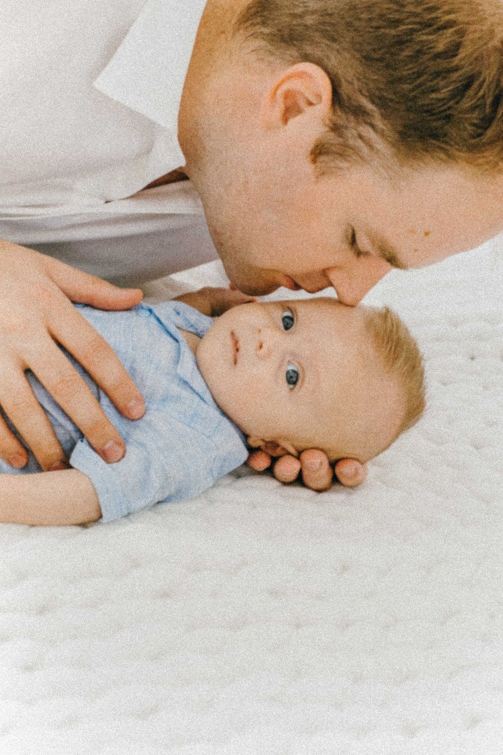Tender moment between parent and baby lying on white bedding. Specializing in maternity, newborn, and family photography