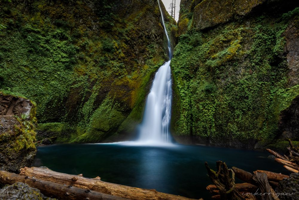 Long exposure waterfall cascades through moss-covered rocks in a lush green forest.