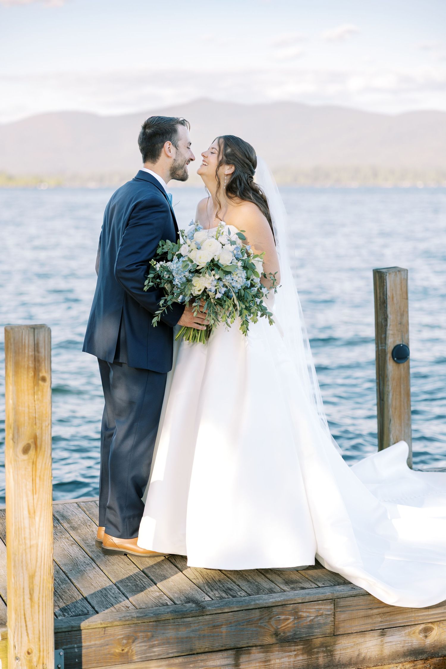 A couple shares a romantic kiss on a lakeside wooden dock during their wedding photoshoot at sunset.