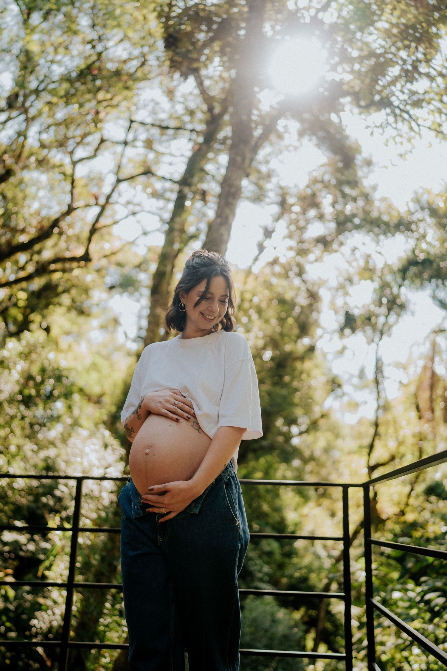 Uma futura mãe vestida de branco está sob a luz do sol entre as árvores.