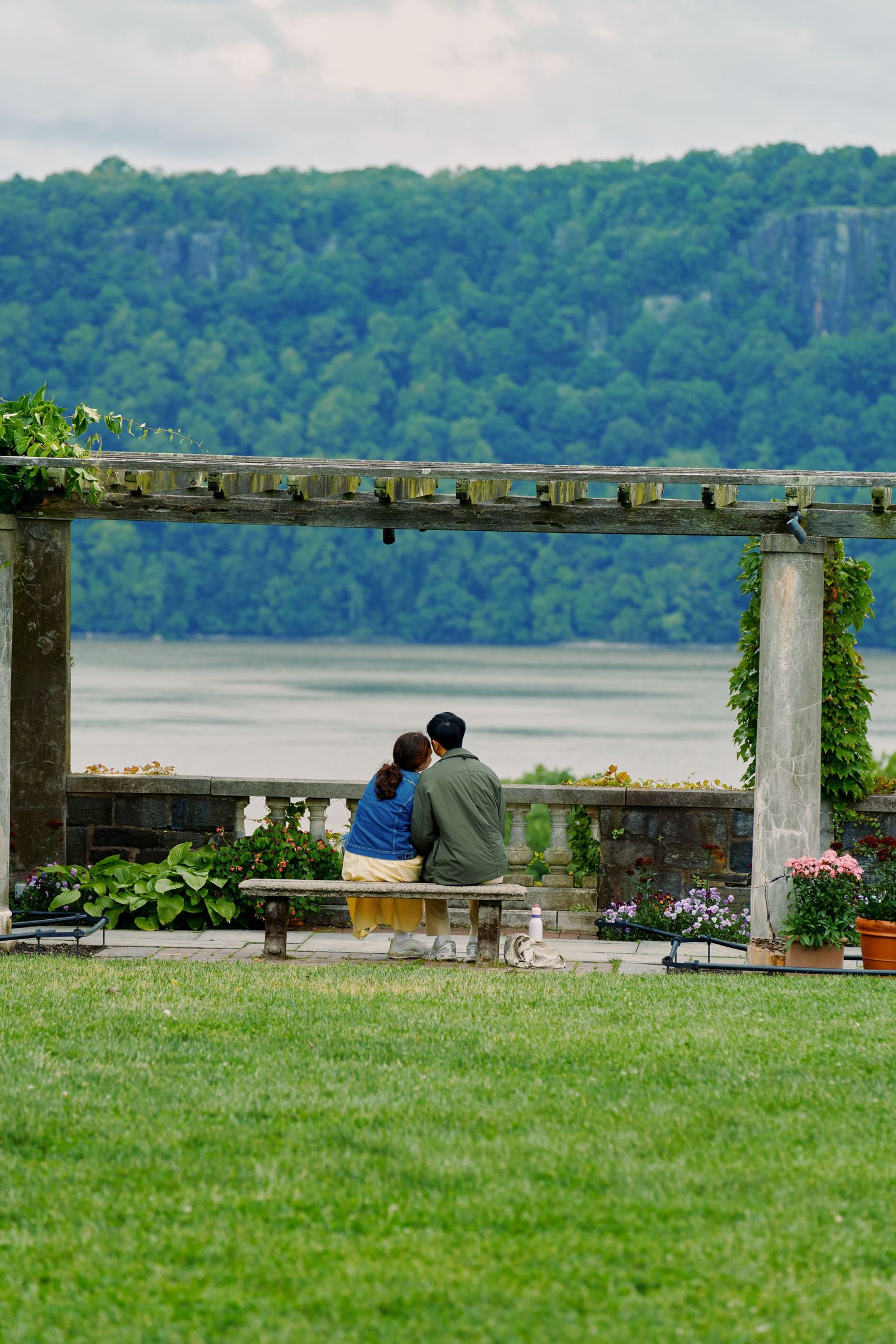 A couple sits together on a bench under a pergola overlooking a scenic lake and forested hillside on a summer day. Wave hill garden