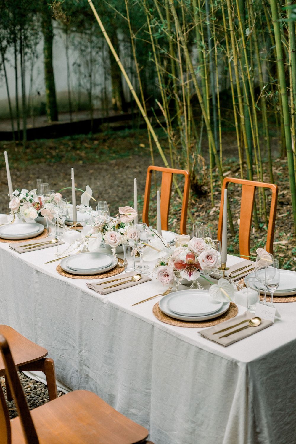 Elegant outdoor table setting with white linens, wooden chairs, and delicate floral arrangements against a bamboo backdrop.