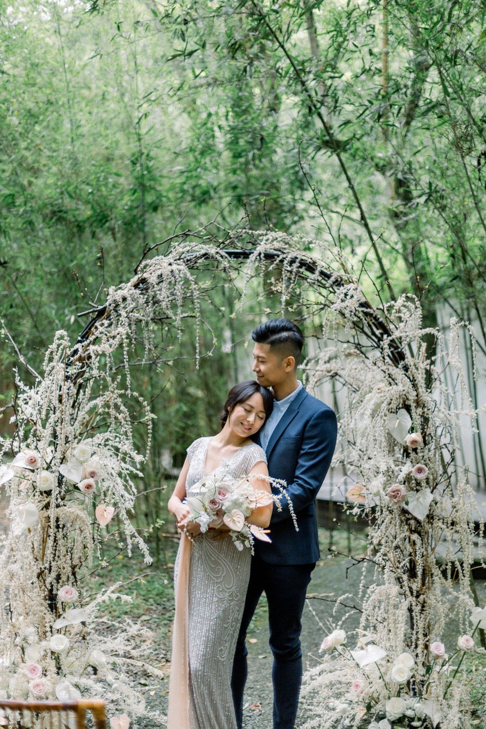 A couple poses under an elegant floral arch made of white flowers in a dreamy forest setting.