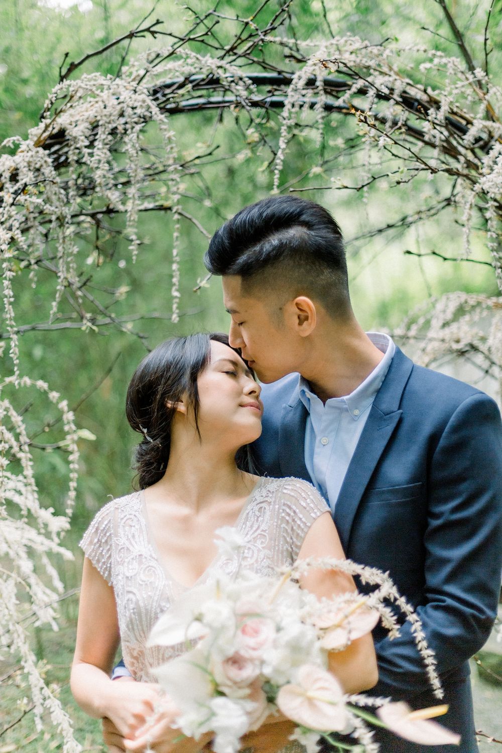 A romantic wedding couple embraces under blooming white cherry blossom branches in a springtime garden setting.