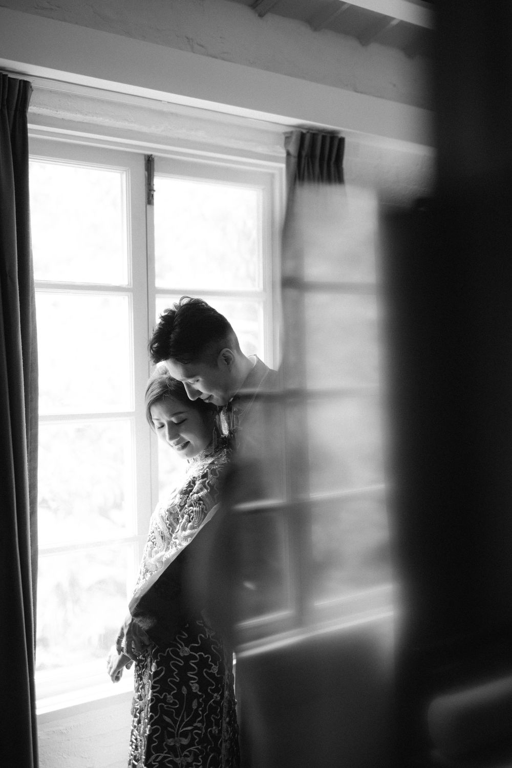 Intimate black and white portrait of a couple embracing by a window with soft natural light streaming in.