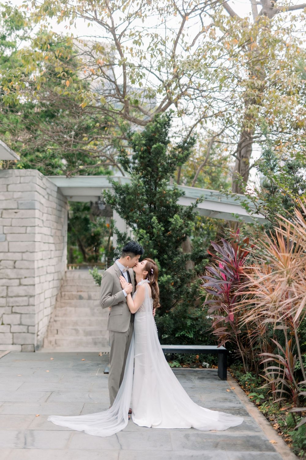 A couple shares an intimate moment in a garden setting with stone pillars and lush greenery surrounding them.
