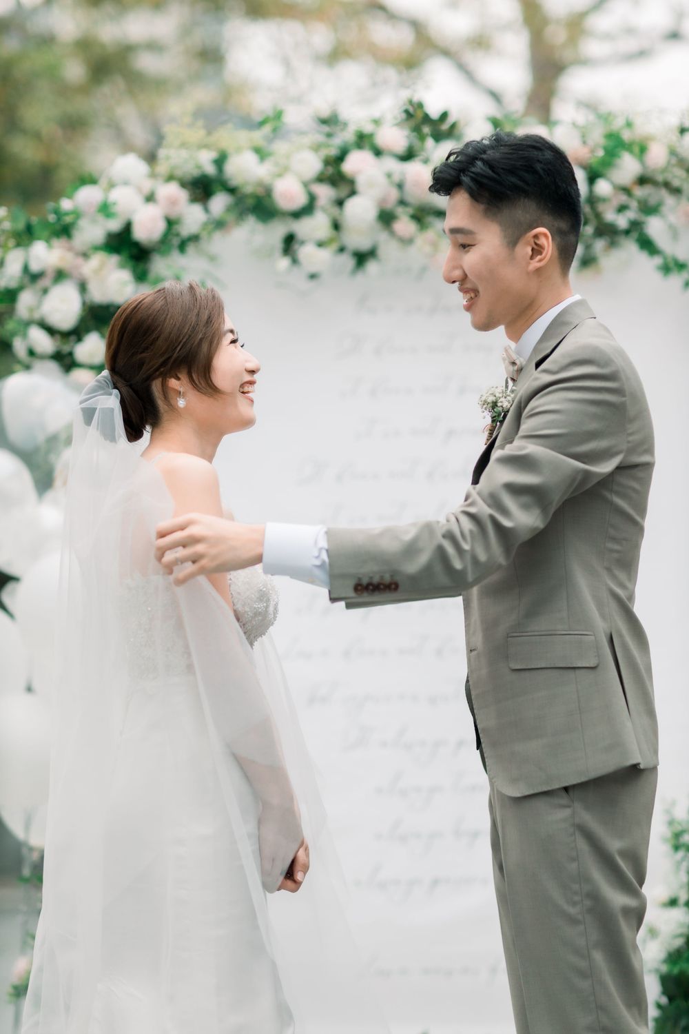 Couple in wedding attire sharing playful moments against a white floral backdrop.