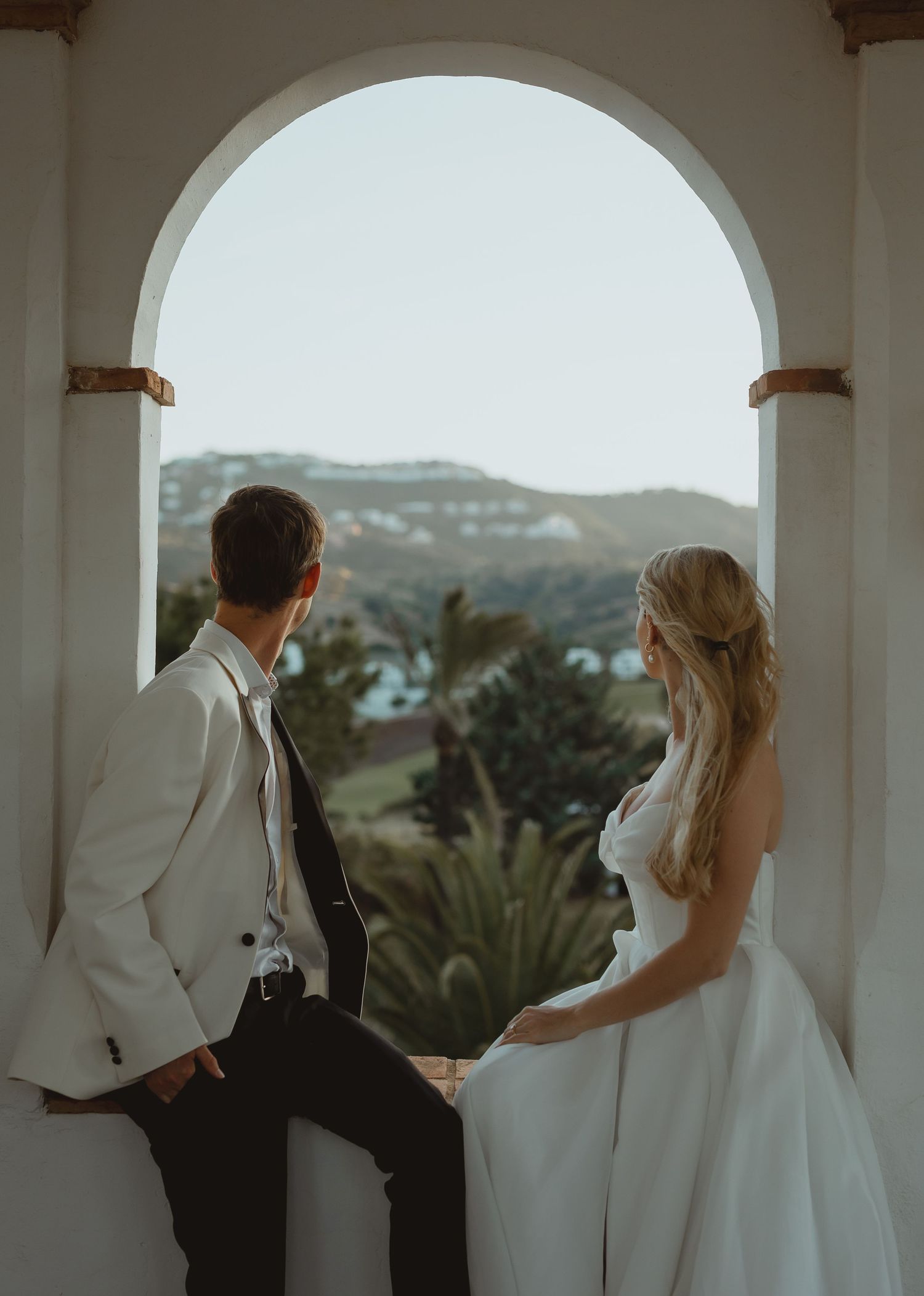 A romantic scene through white arches shows a couple in wedding attire looking out at a scenic mountain landscape.