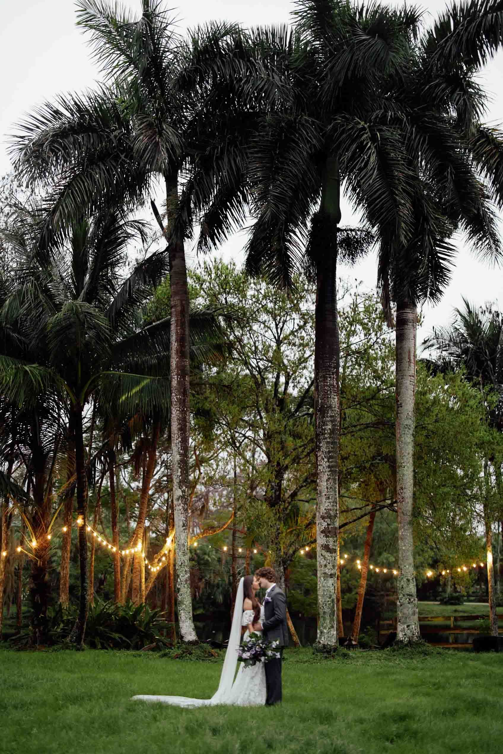A romantic outdoor wedding scene under palm trees with string lights at sunset in a lush garden setting.