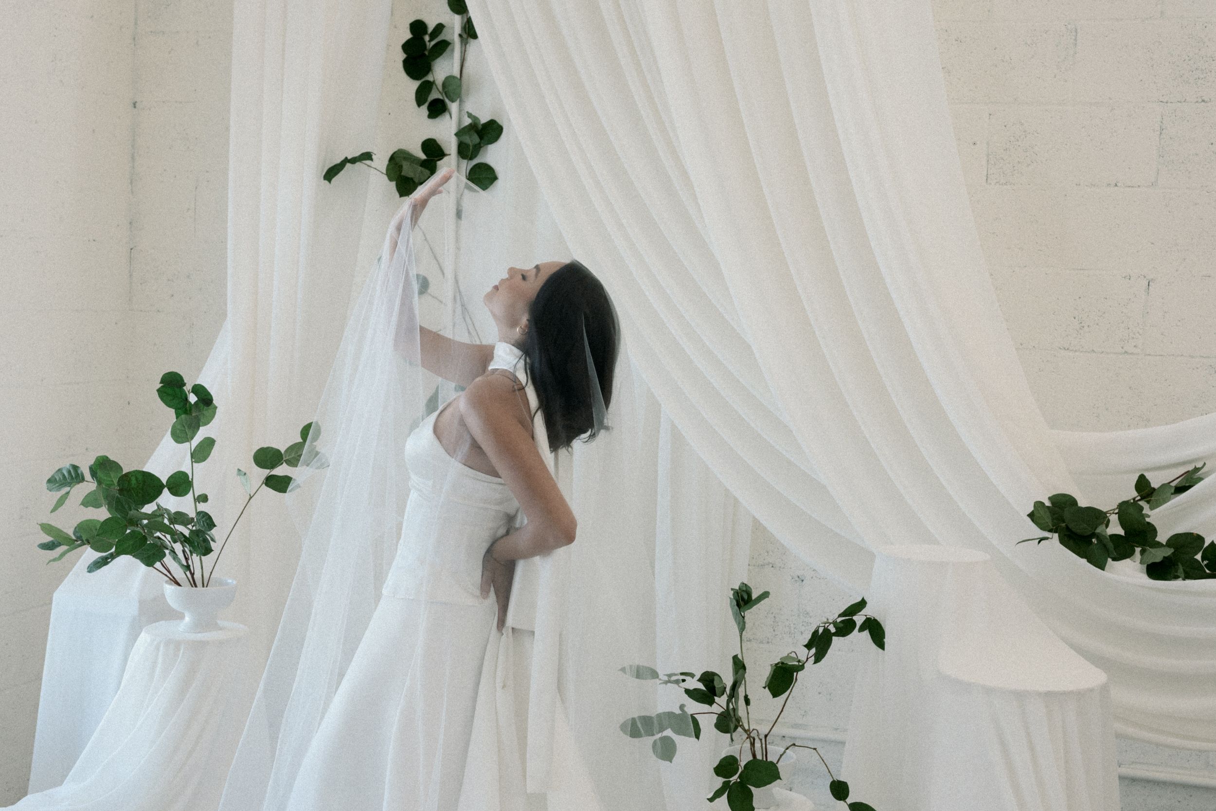 A white wedding dress hangs elegantly against a backdrop of cascading white curtains and delicate green vines.