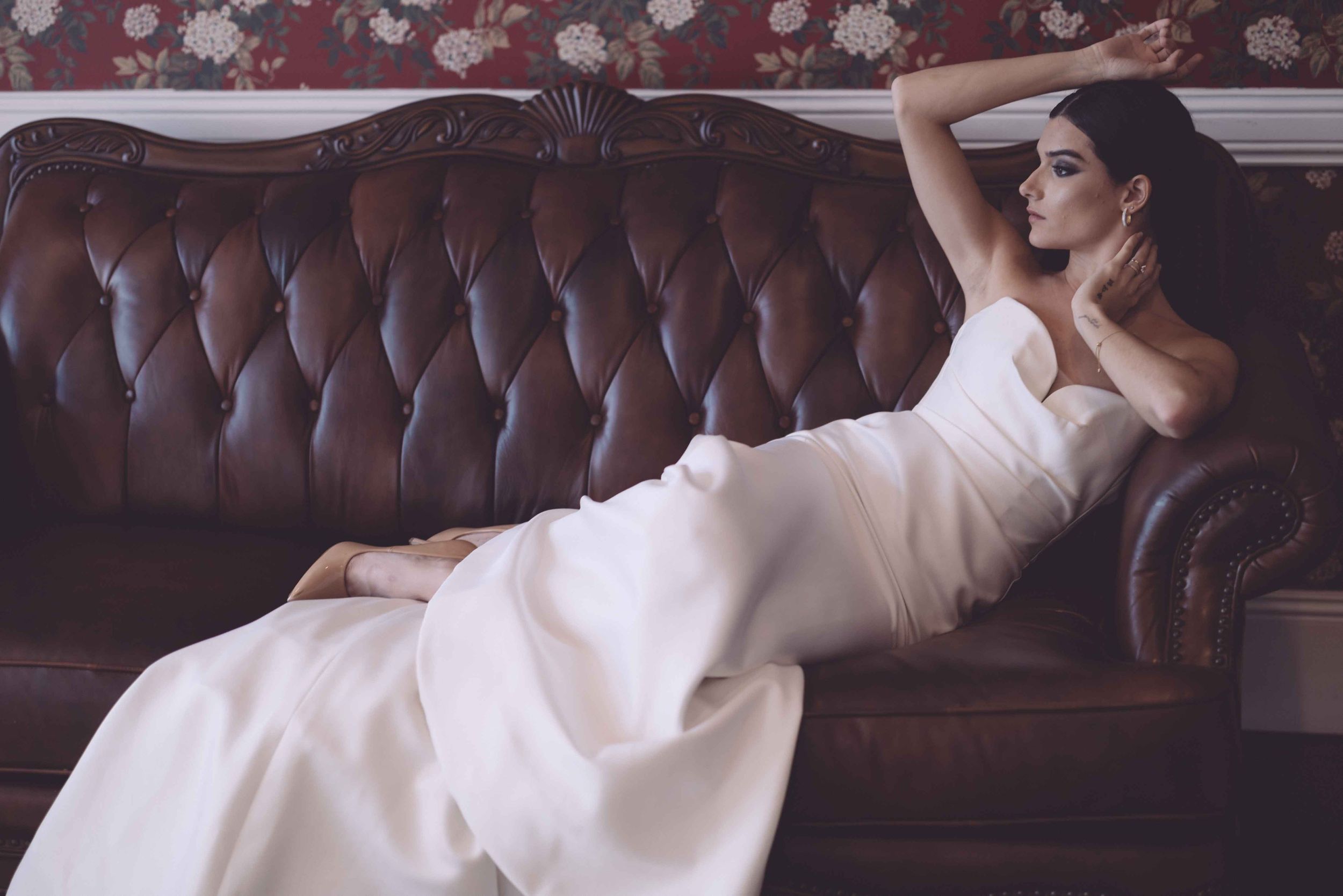 Ballet dancer in white flowing dress performs graceful poses against dark leather couch backdrop.
