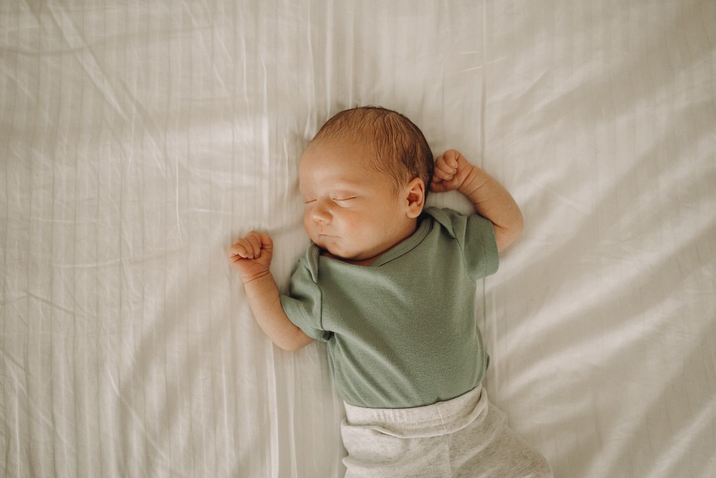 Infant resting on white bedding wearing sage green clothing.