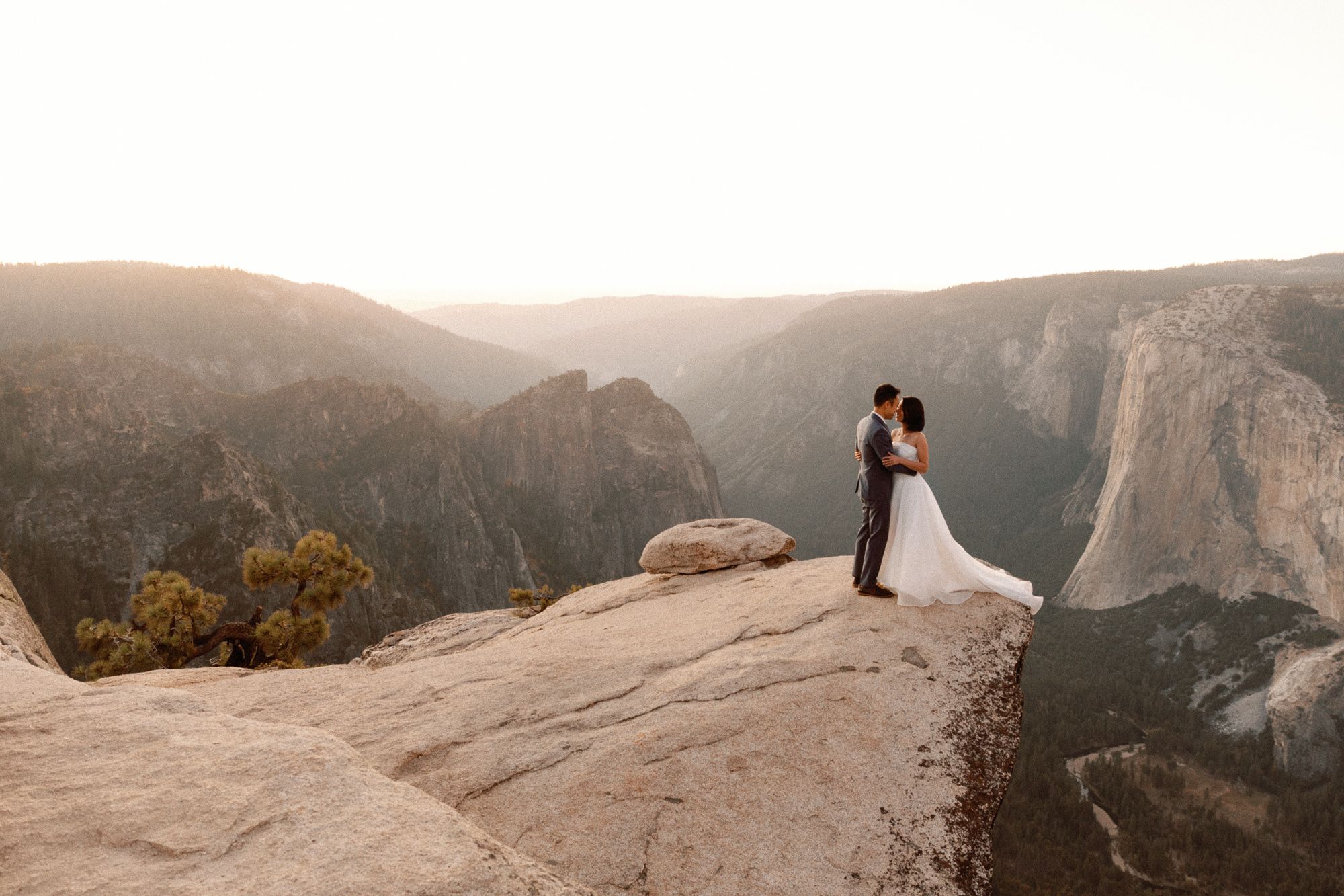 Bride and groom kiss on rocky Yosemite cliff edge at golden sunset with valley views.