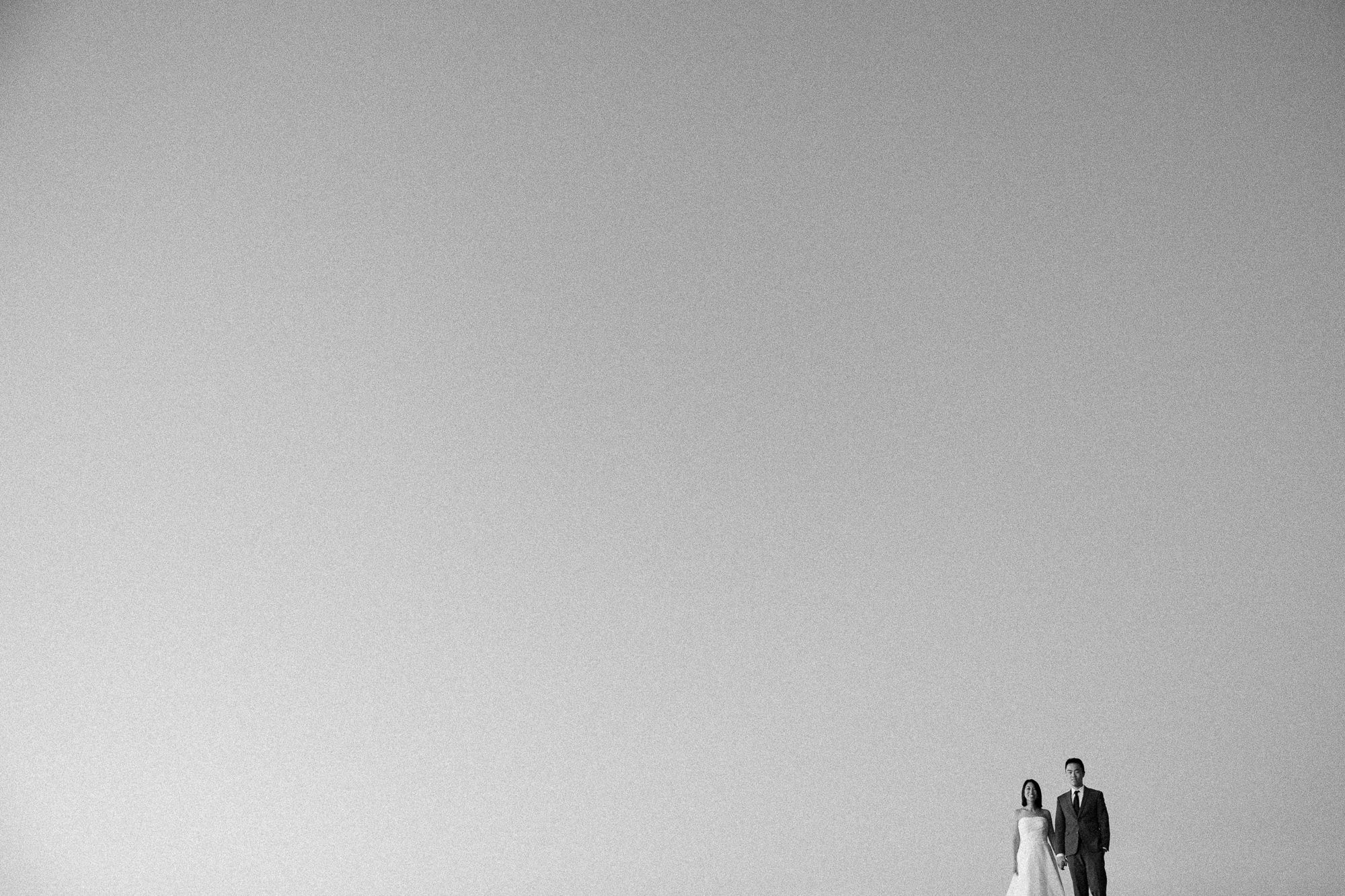 Couple stands together in minimalist black and white wedding photo against vast open sky.