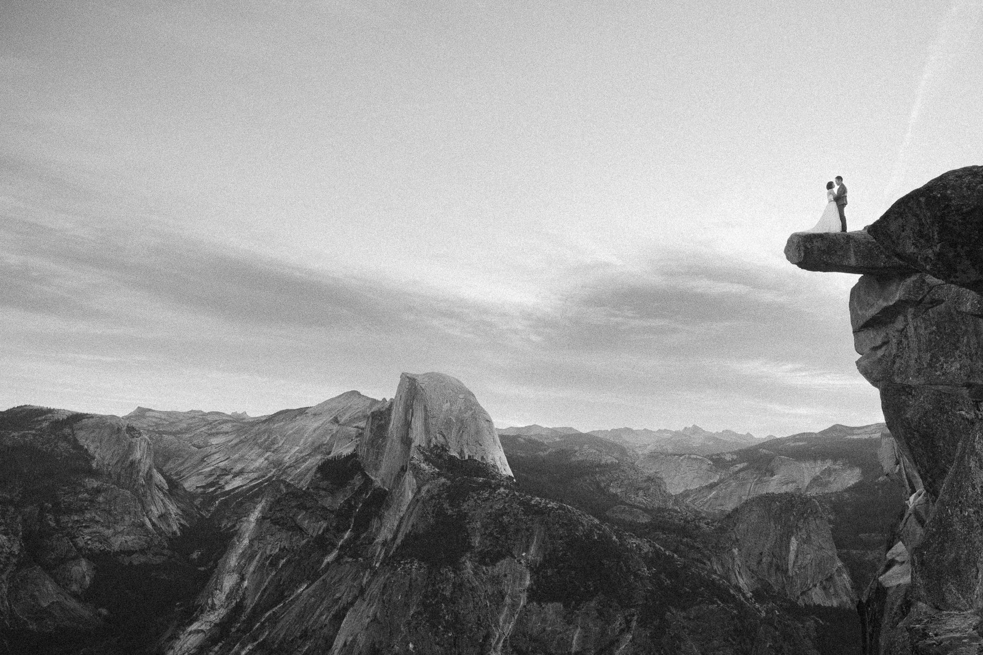 Black and white photo of couple standing on Yosemite cliff edge overlooking Half Dome.
