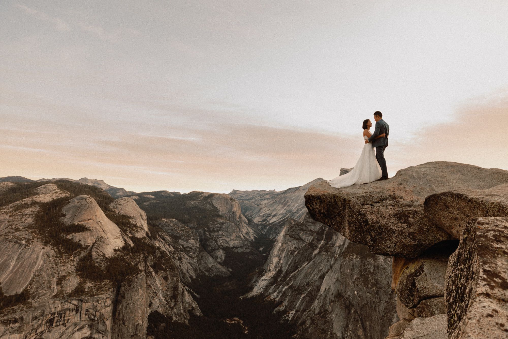 Wedding couple embraces on rocky Yosemite overlook with Half Dome visible in background.