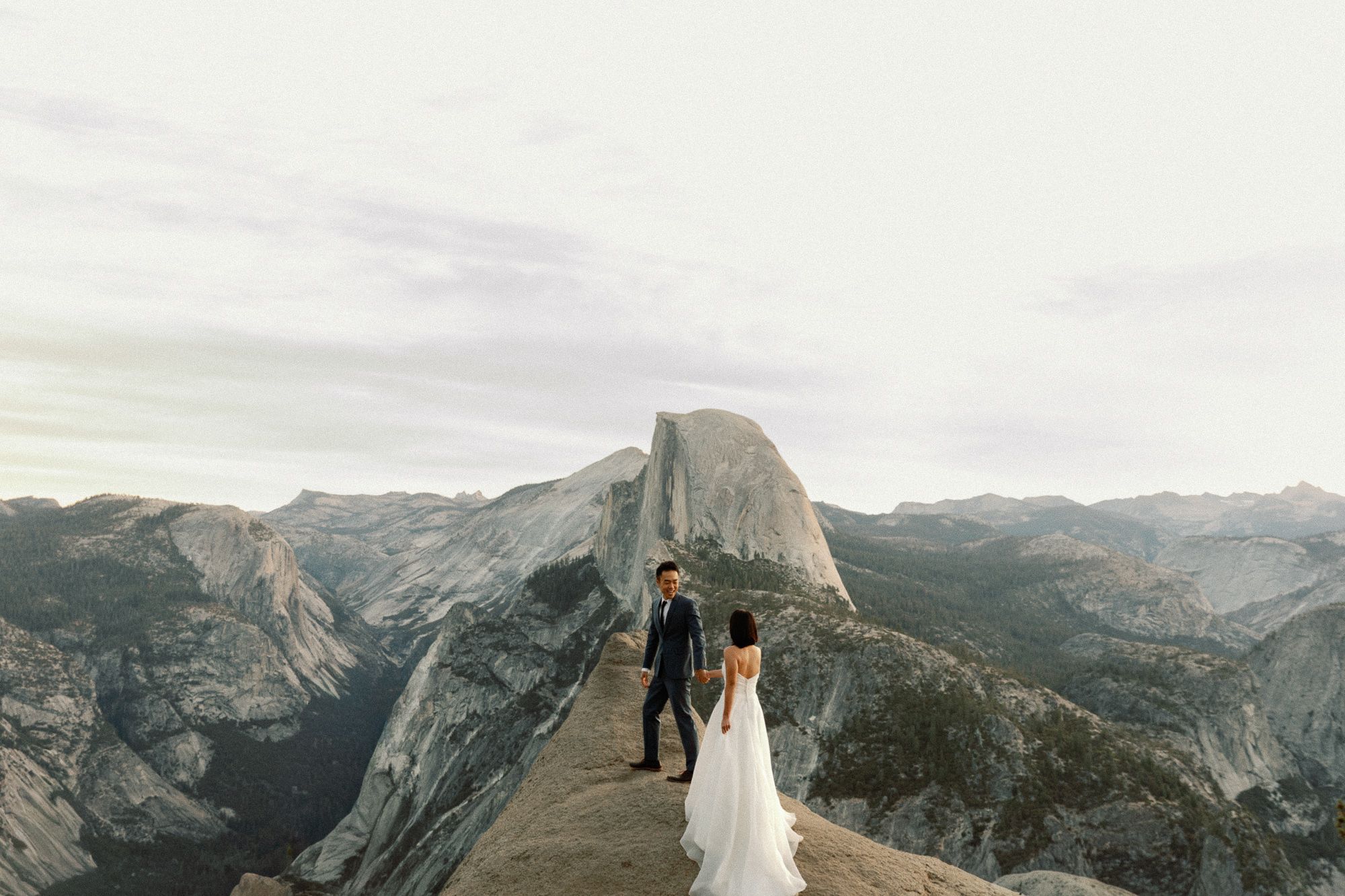 Bride and groom hold hands at Glacier Point with Half Dome and Yosemite Valley behind them.