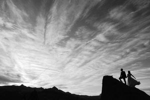 Dramatic black and white silhouette of bride and groom on rocky peak under wispy clouds.