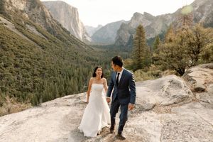 Smiling bride and groom walk hand in hand on granite overlook above Yosemite Valley.