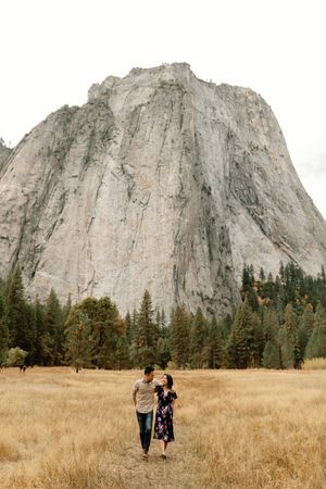 Couple walks through golden meadow with massive granite cliff face rising behind them.