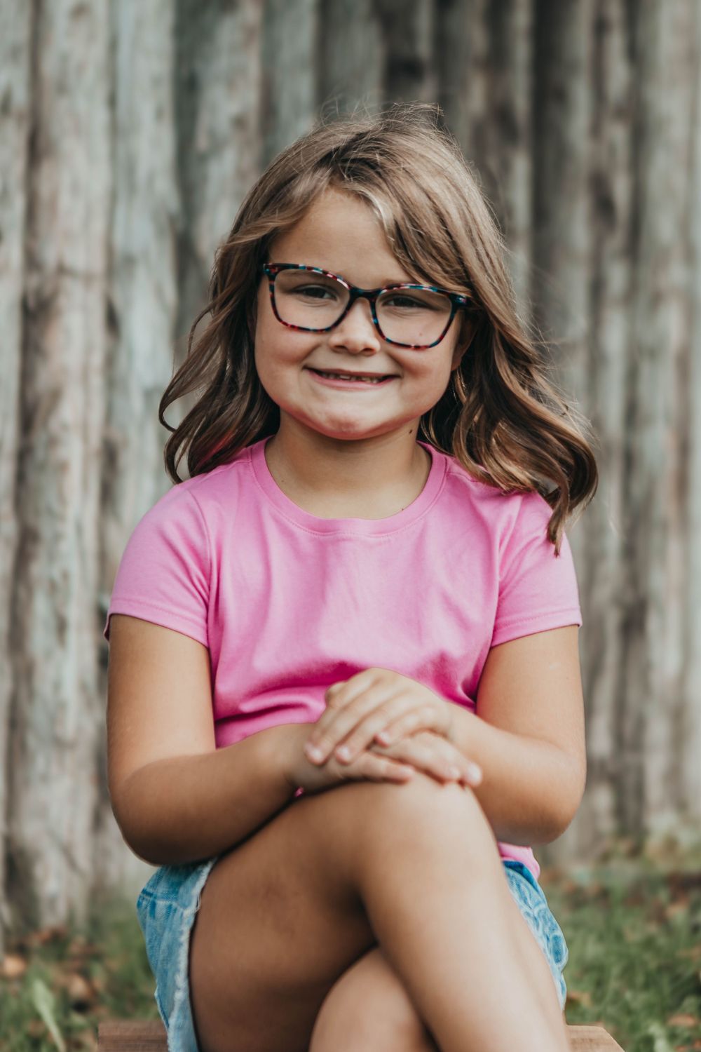A young child in a pink shirt and glasses sits casually on grass against a wooden fence.