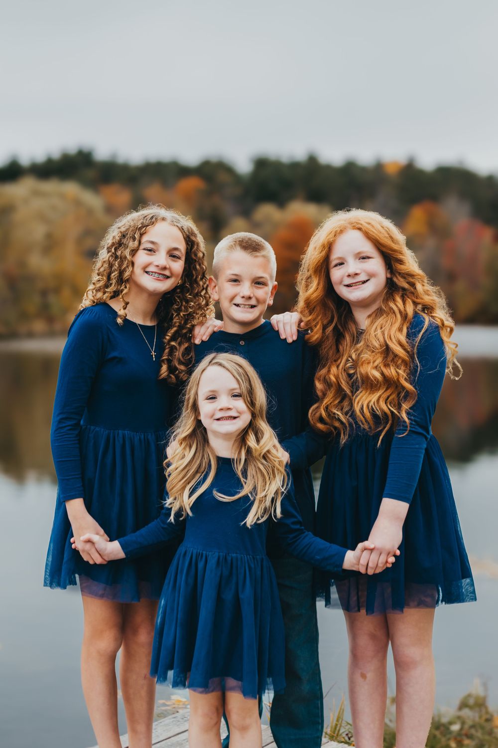 Group of children in matching navy blue dresses pose by a lake during autumn.