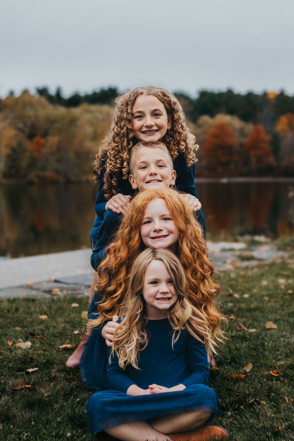 Three siblings in matching navy blue outfits pose playfully near a lake during autumn.