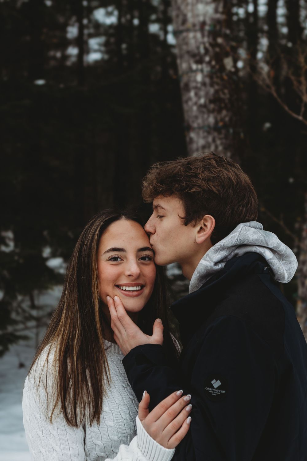 Couple shares intimate winter moments in the snow wearing cozy sweaters among pine trees.