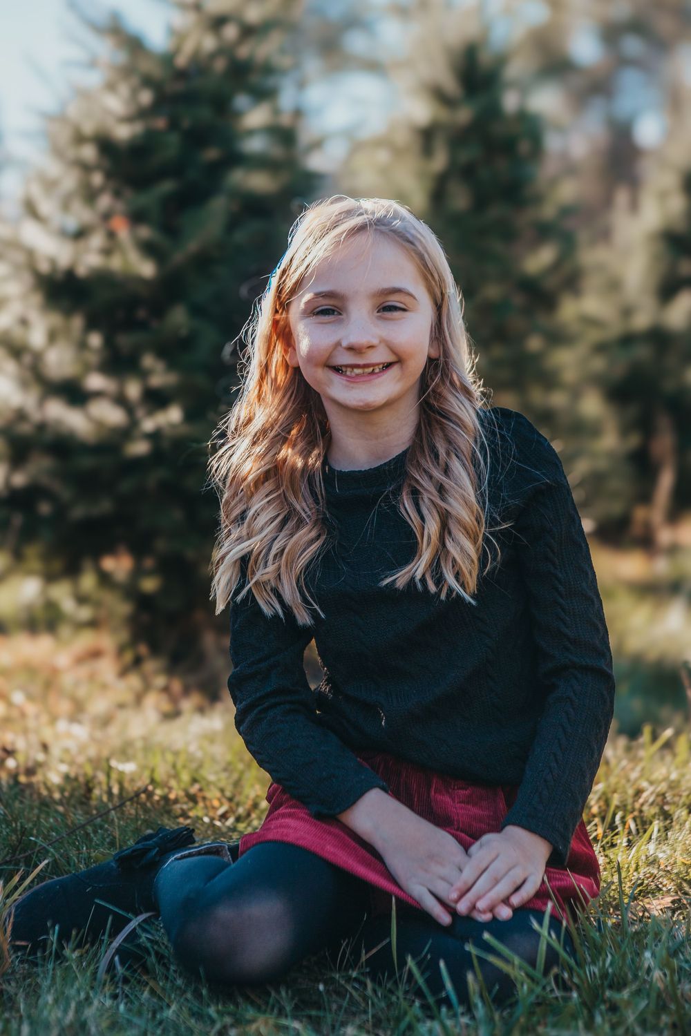 A person in a black sweater sits in grass during golden hour with evergreen trees in the background.