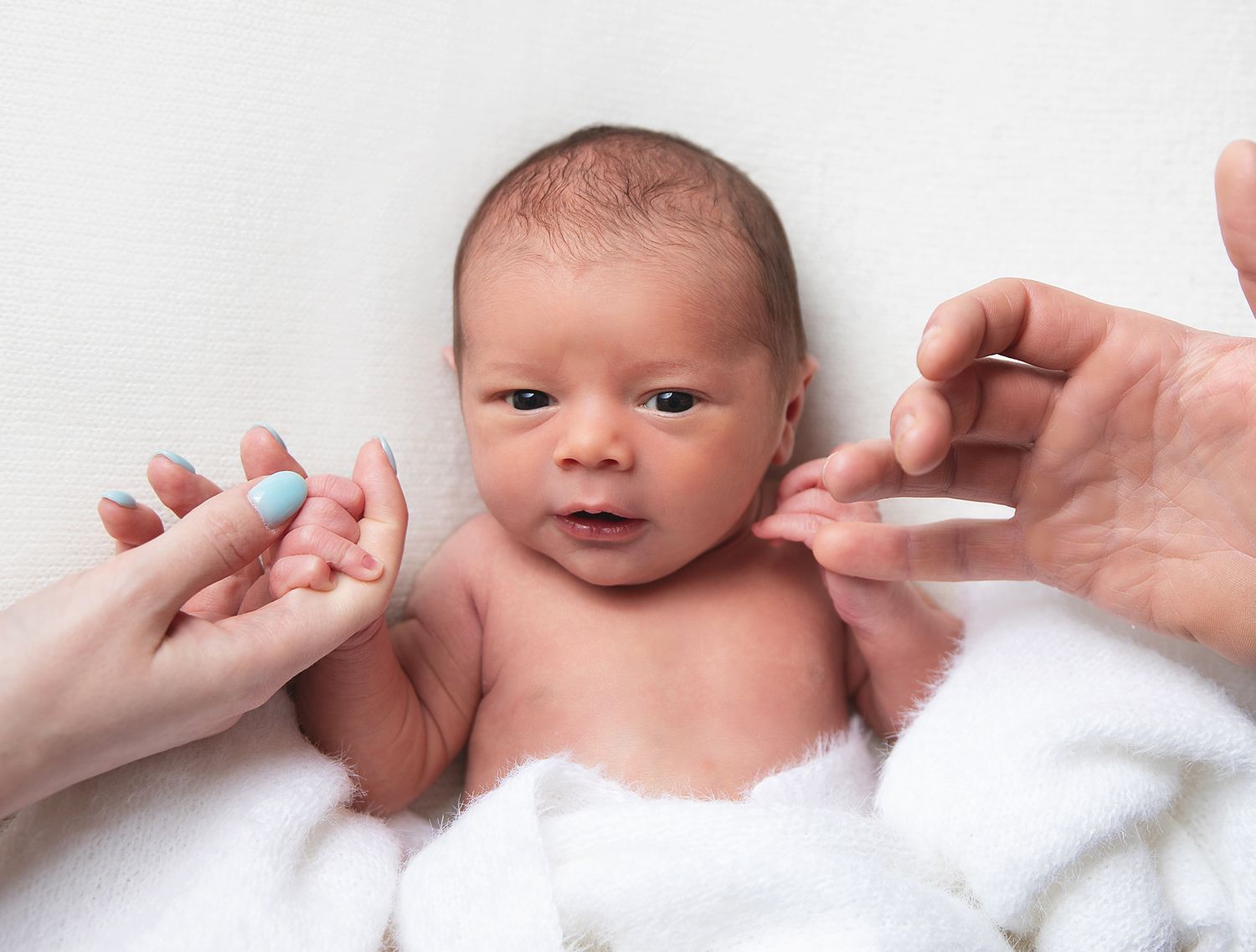 Newborn baby wrapped in white blanket while hands gently touch their face during medical checkup. During a newborn photoshoot in Sidcup