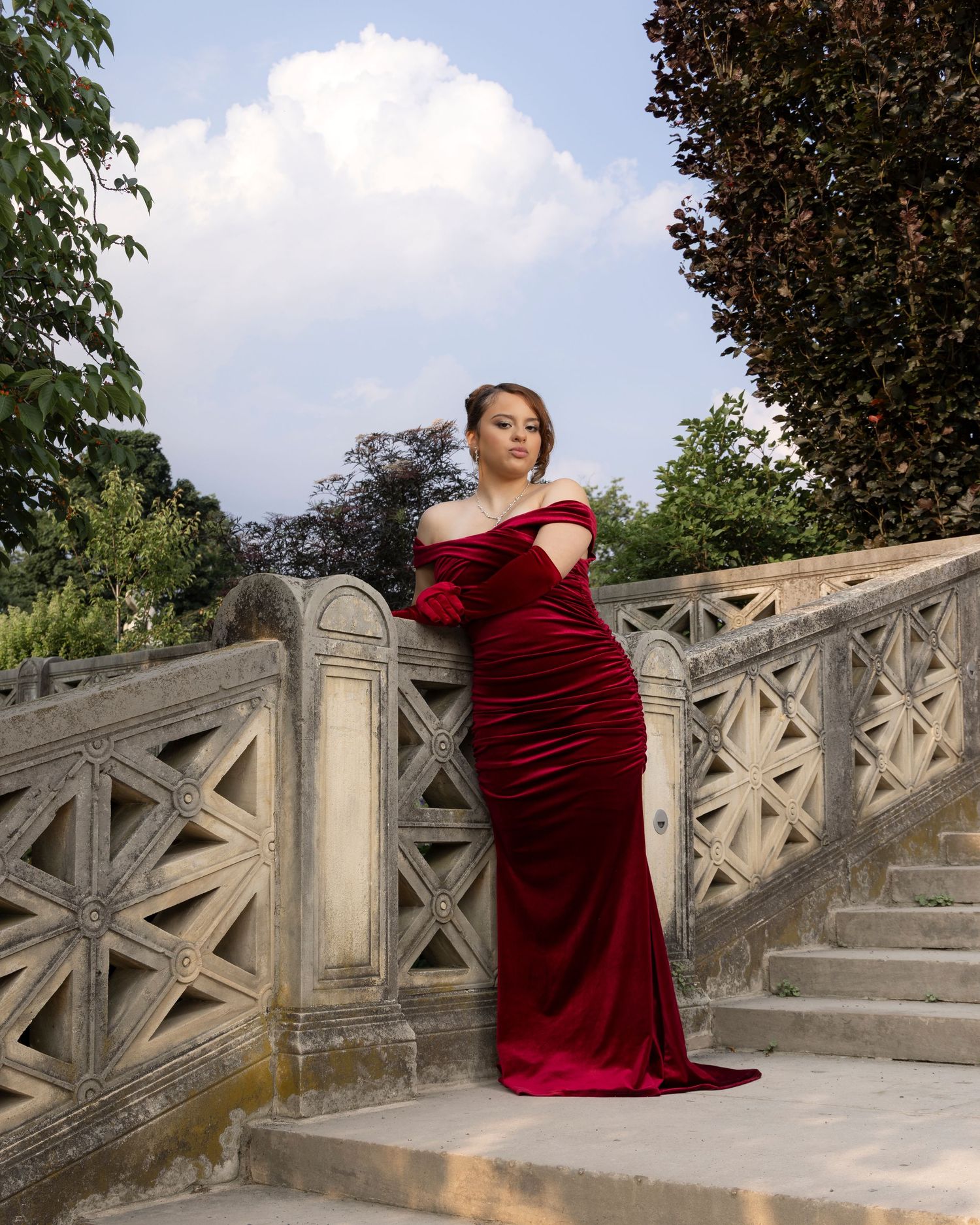 Elegant figure poses on concrete steps in a flowing burgundy velvet evening gown amidst lush garden scenery.