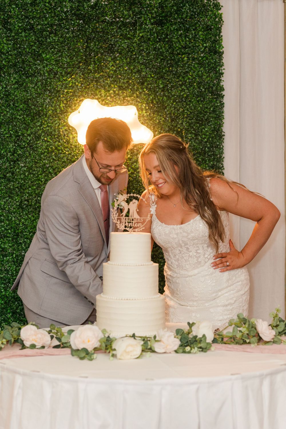 Wedding couple blowing out candles together from multiple angles on their white tiered cake with greenery backdrop.