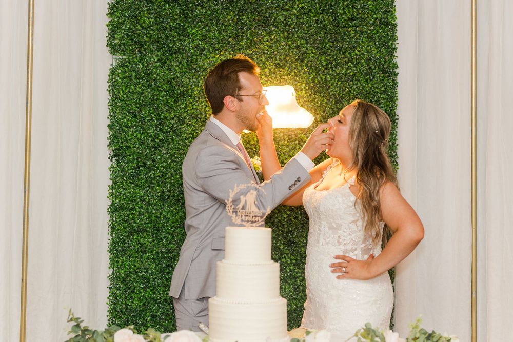Newlyweds share cake by their white wedding cake against a green hedge wall with glowing lighting.