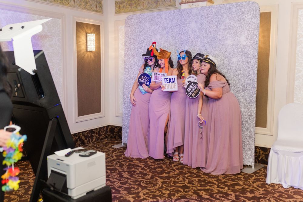 People in matching lavender bridesmaid dresses pose together at a photo booth with a glittery silver backdrop.