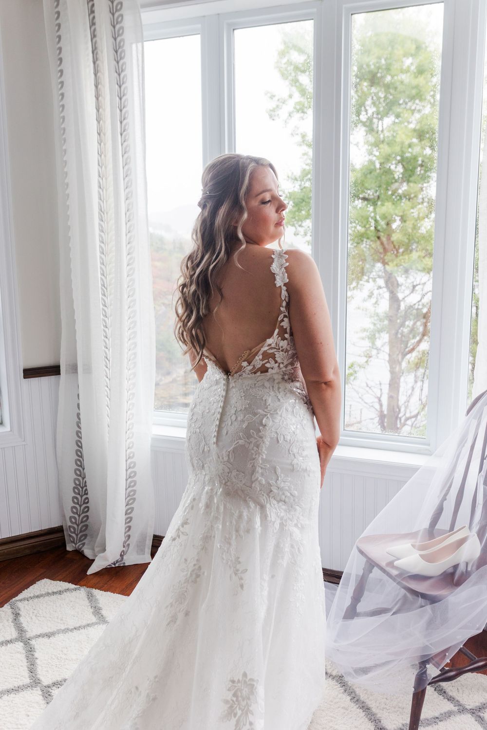 Bride in lace detailed white wedding gown with long sheer veil poses gracefully near sunlit windows.