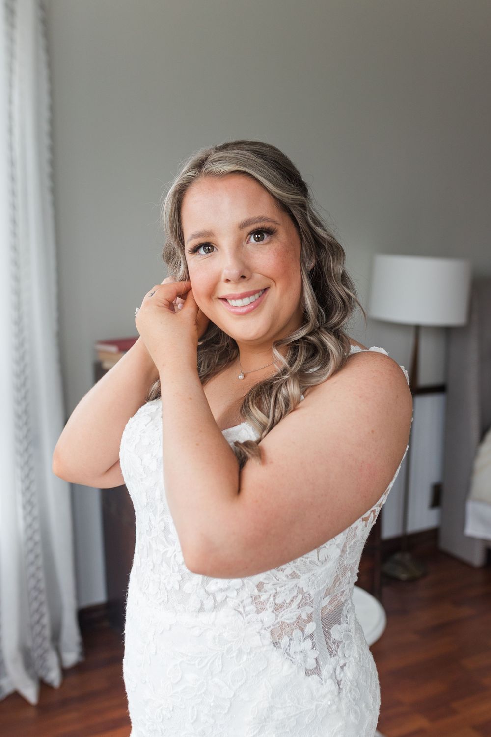 A bride wearing a white lace wedding dress poses against light gray walls in a room with hardwood floors.