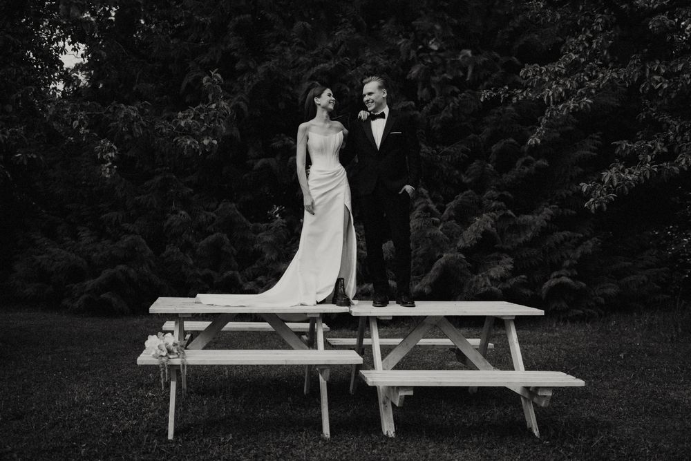 A couple in wedding attire stands together on a picnic table in a garden setting.