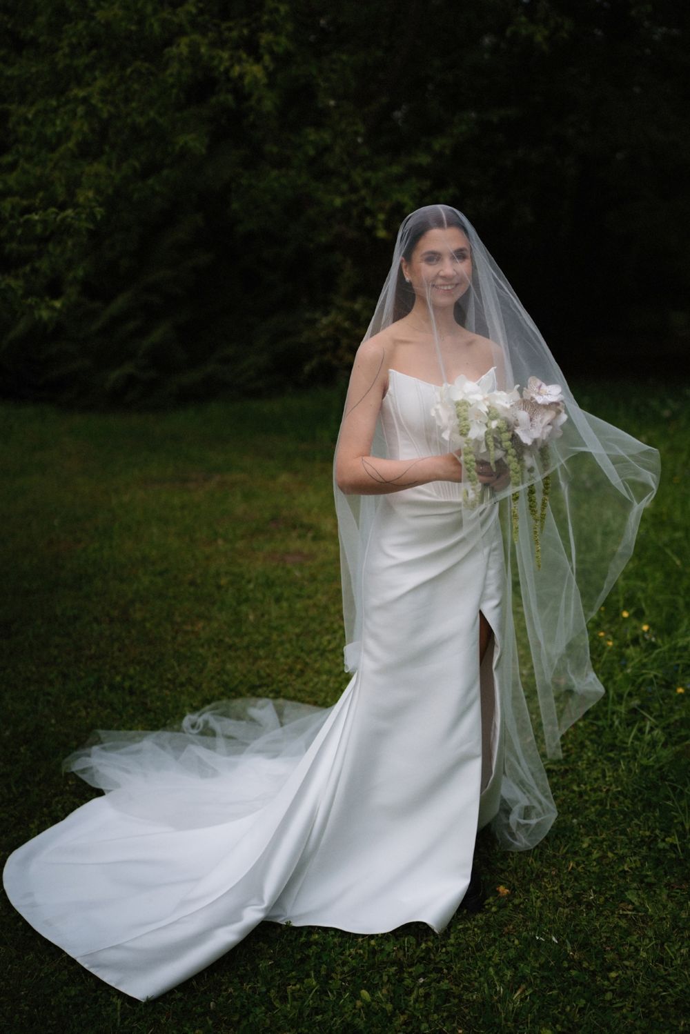 Elegant bride in flowing white dress and veil holding bouquet in garden setting.