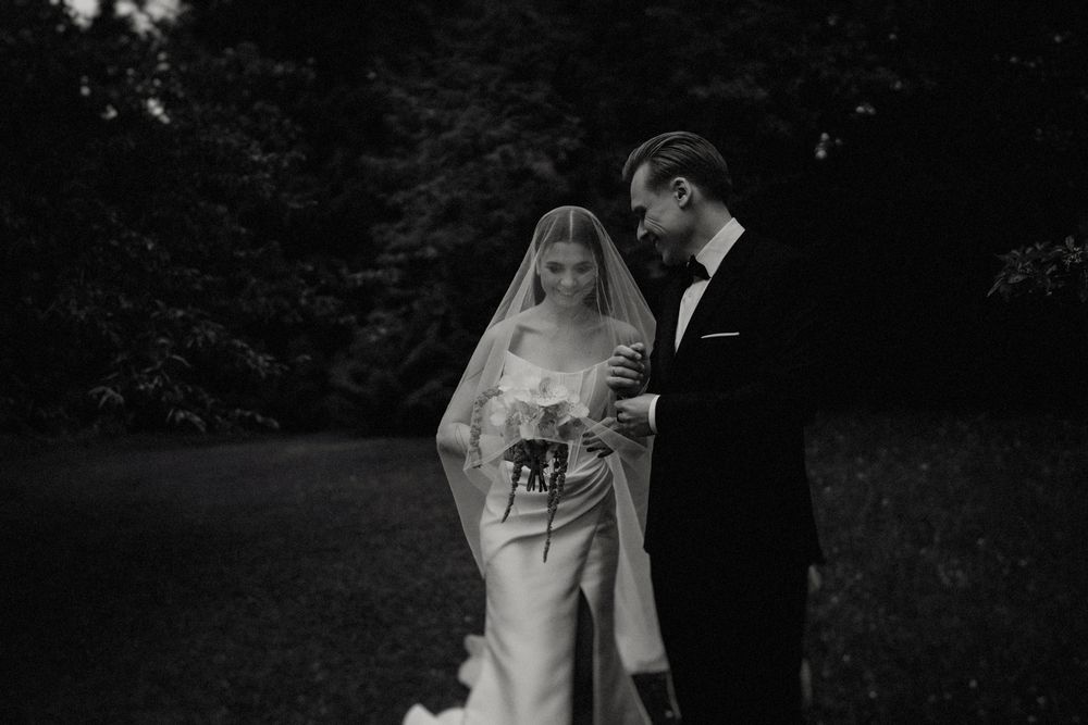 Black and white wedding photo in garden setting with elegant attire and bouquet.