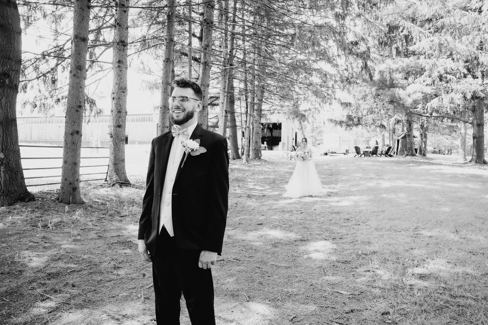 A figure stands among tall evergreen trees in a wooded park setting wearing formal black attire.
