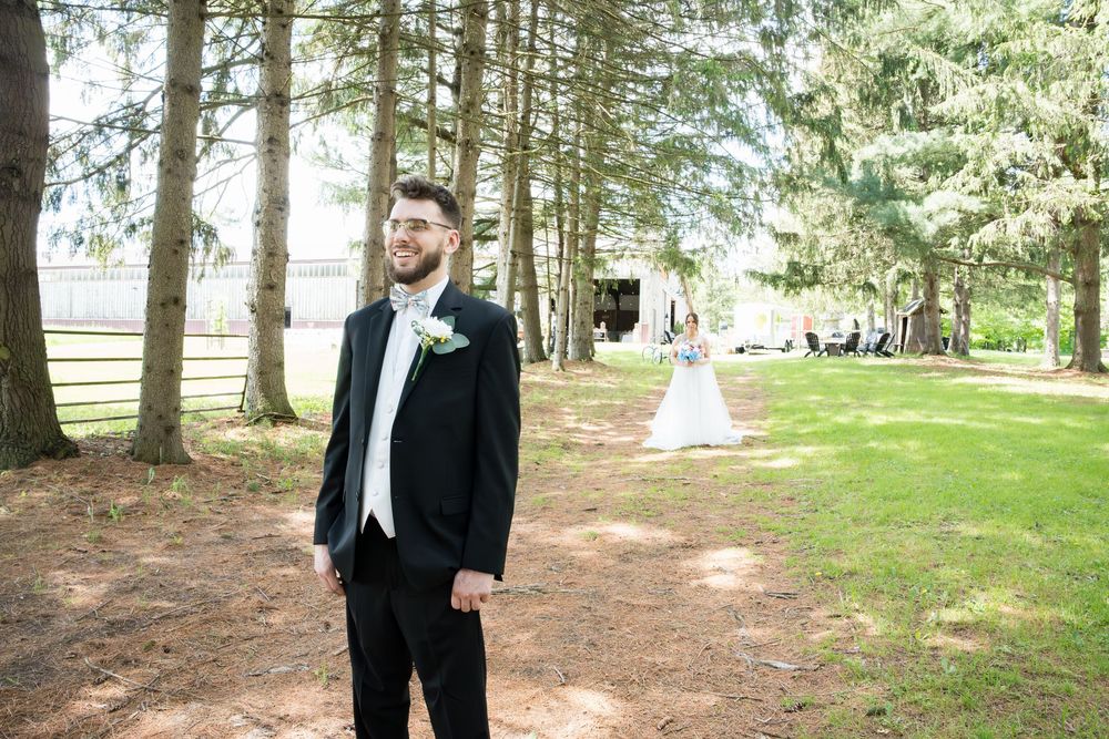 Figure in a classic black tuxedo poses elegantly against a backdrop of sunlit pine trees in an outdoor setting.