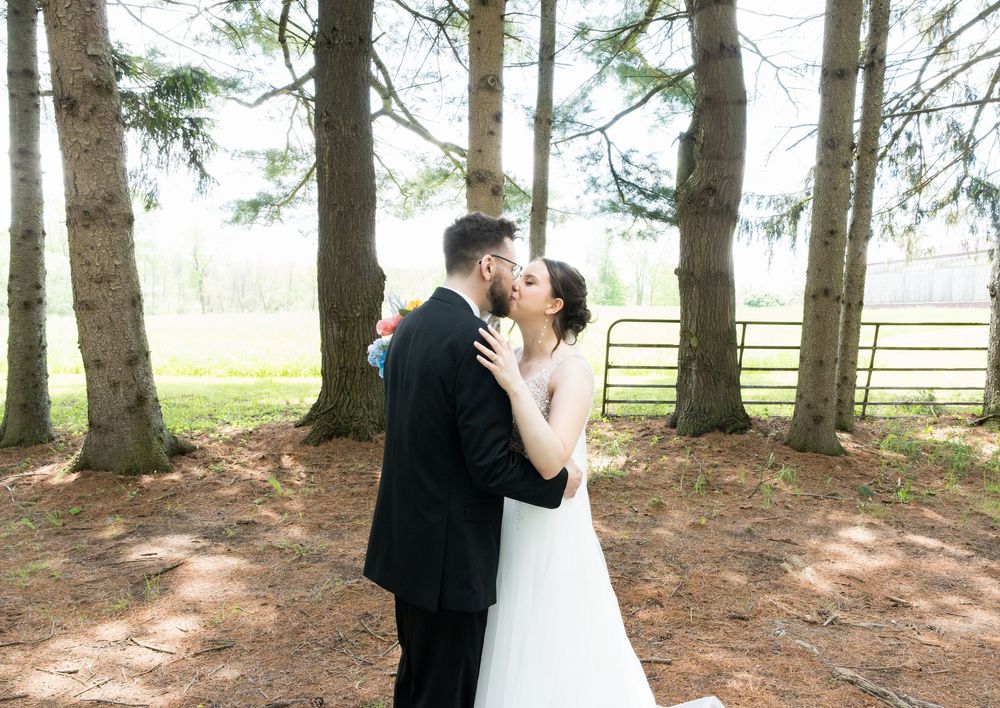 A couple shares a romantic kiss under tall evergreen trees on their wedding day at a rustic farm location.