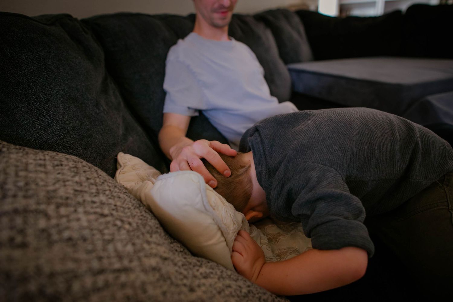 Two people relaxing together on a dark couch in a dimly lit living room setting.