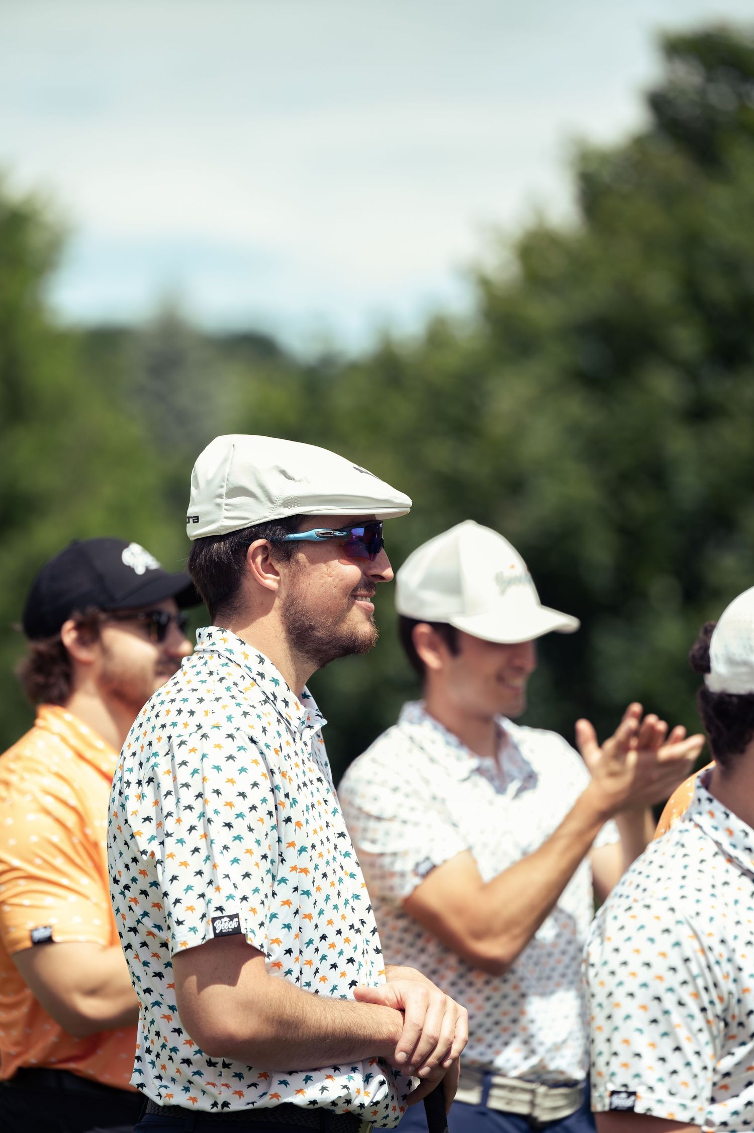 Golfers in casual attire with flat caps and patterned shirts stand together on the golf course during a sunny day.